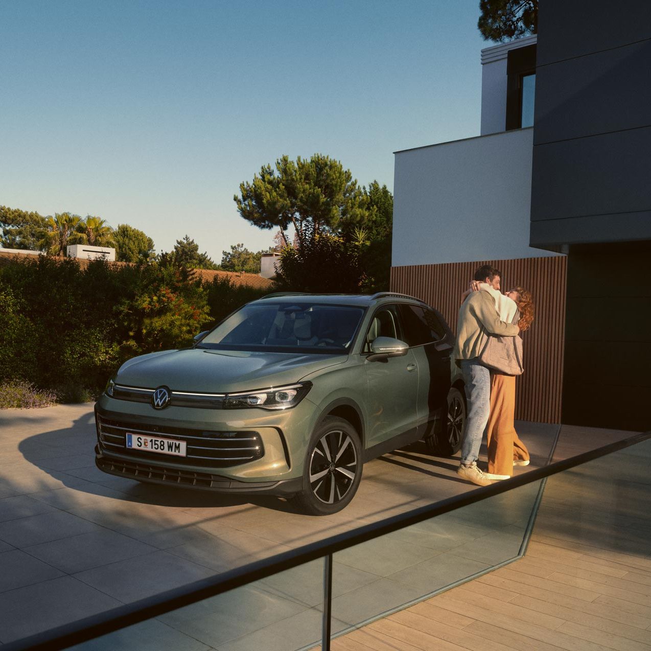 Grüner VW Tiguan parkt in Frontansicht auf der Einfahrt eines modernen Wohnhauses. Ein junges Paar steht daneben und umarmt sich.