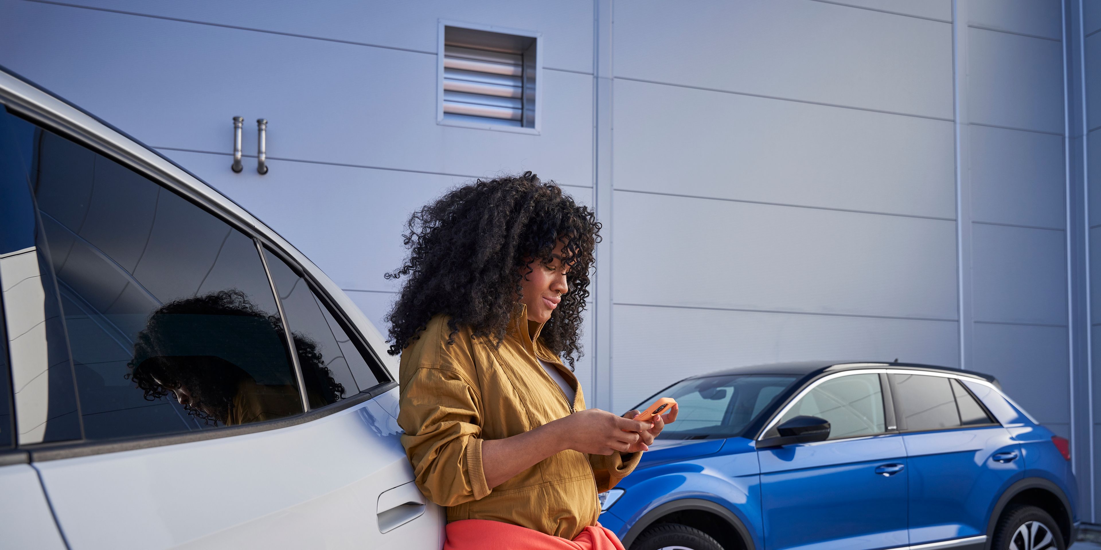 Frau lehnt an Volkswagen und blickt auf ihr Smartphone, im Hintergrund sieht man einen blauen T-Roc