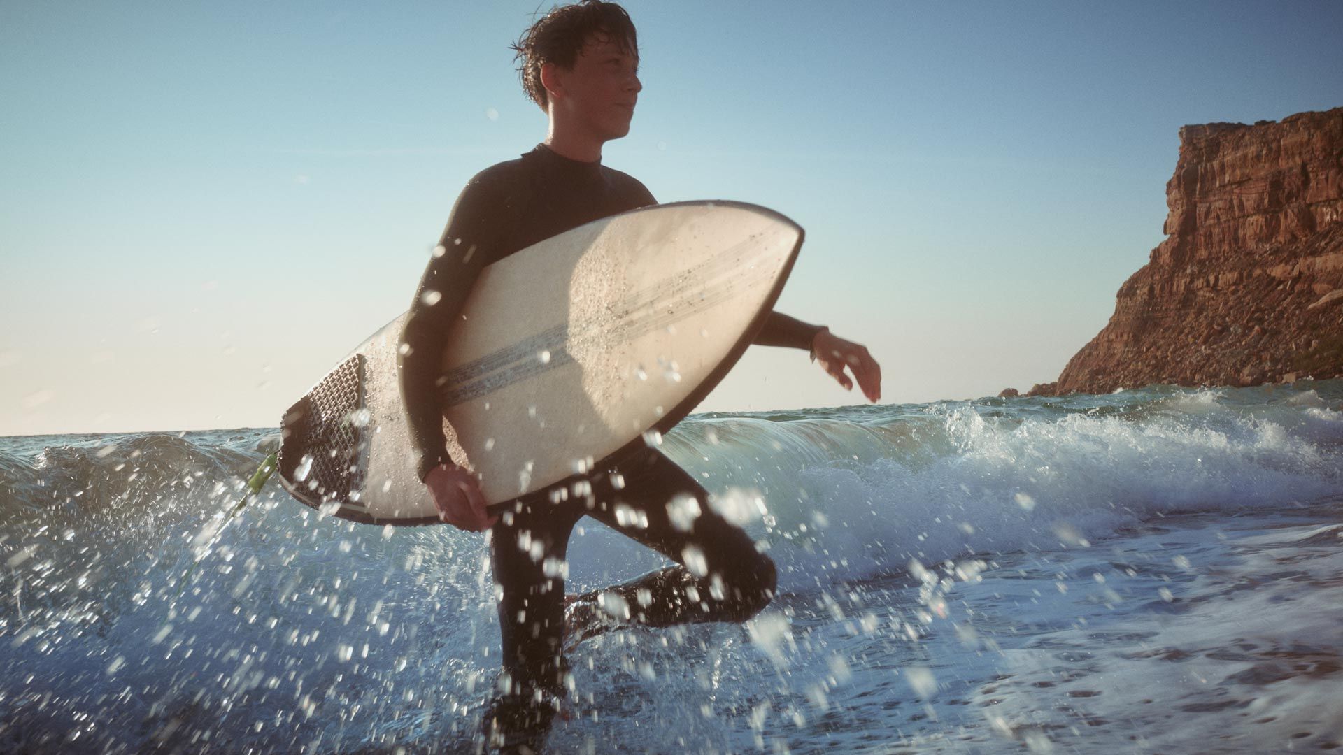 Scenă la plajă - un tânăr merge prin apa adâncă până la genunchi cu o placă de surfing la subraț.  