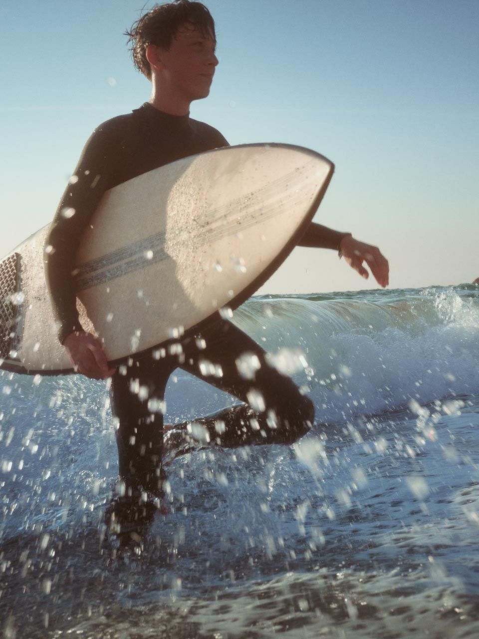 Scenă la plajă - un tânăr merge prin apa adâncă până la genunchi cu o placă de surfing la subraț.  