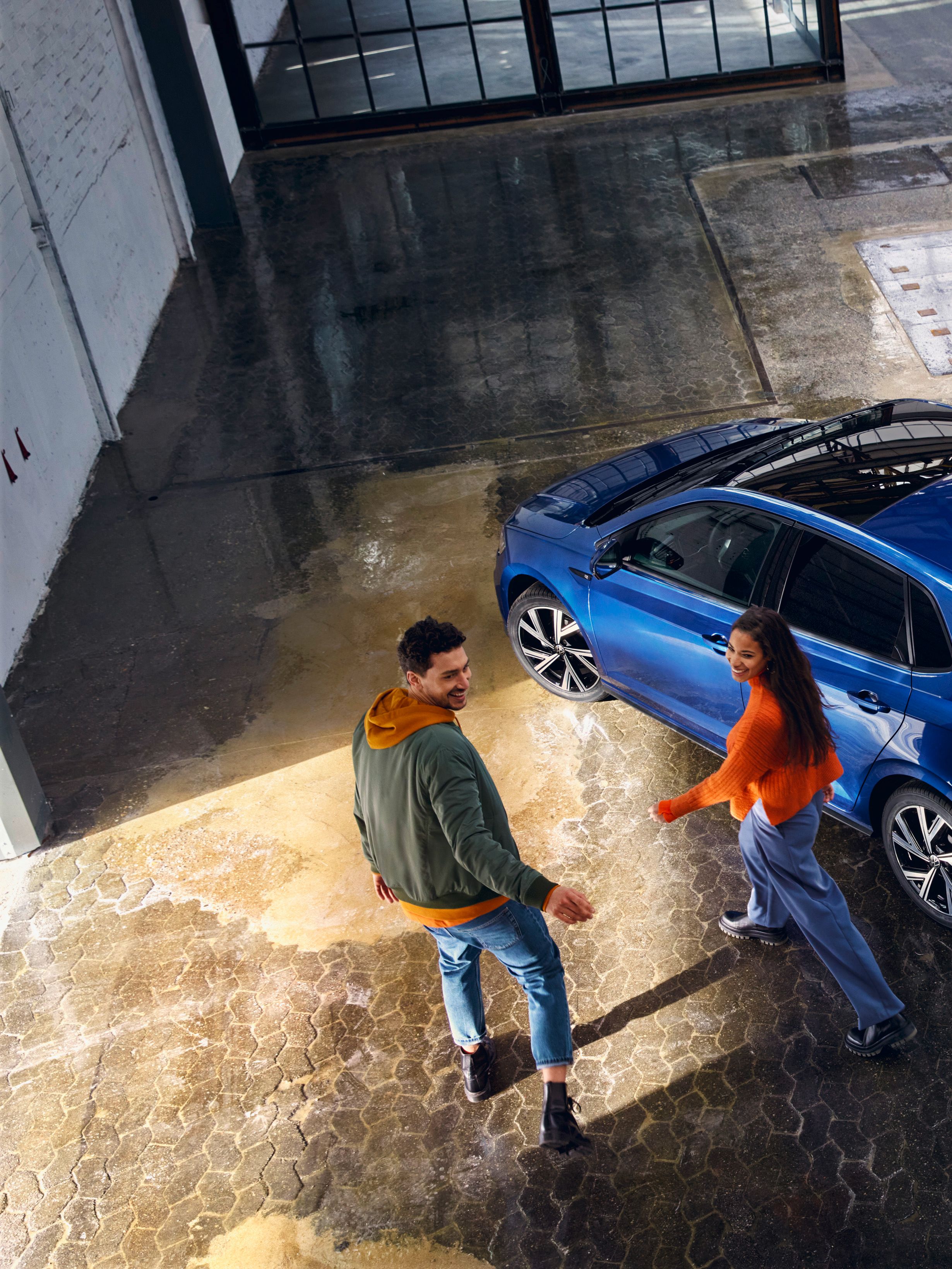 Ein VW Polo in Blau mit optionalem Panoramaschiebedach steht in einer Halle, ein Pärchen geht darauf zu.