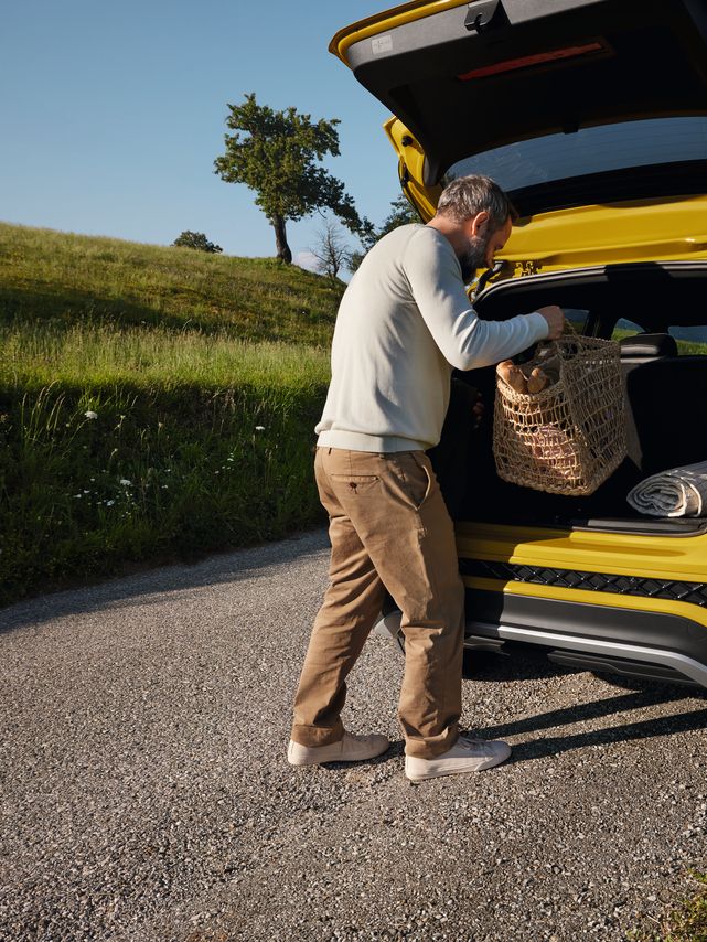 Un bărbat așază un coș de picnic mare în portbagajului unui VW T-Cross galben, aflat pe un drum într-un peisaj cu dealuri.